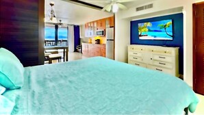 Une chambre avec un lit, un ventilateur de plafond, un téléviseur fixé au mur et une vue sur la cuisine et le balcon.