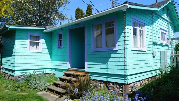 Een turquoise houten huis met een veranda, een kleine tuin met blauwe bloemen en een fundering van natuursteen.