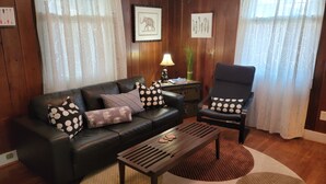 Een woonkamer met een zwarte leren bank, een houten salontafel en een zwarte fauteuil. Er zijn kamergordijnen met een patroon, een lamp en lijstwerken aan de muur.