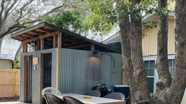 A covered outdoor patio with a table and chairs, surrounded by trees and a corrugated metal wall.