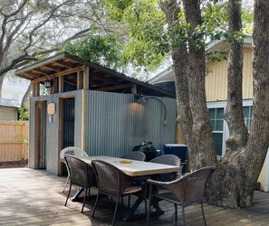 Un patio techado al aire libre con una mesa y sillas, rodeado de árboles y un muro de lámina corrugada.