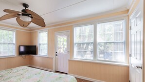 A bedroom with a bed, a ceiling fan, a TV, and large windows with blinds.