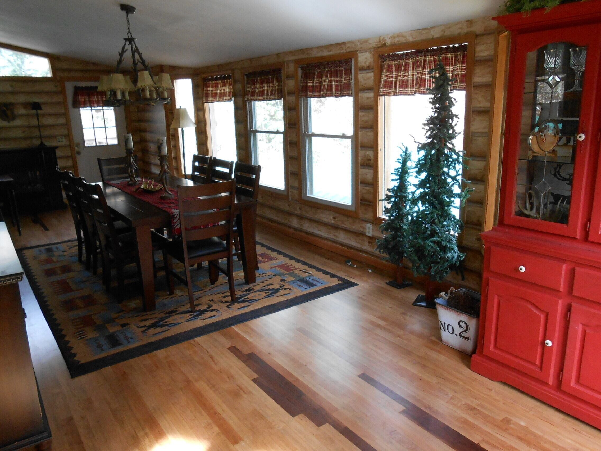 A log cabin interior with a dining area, wooden floors, plaid window valances, a Christmas tree, and a red cabinet.