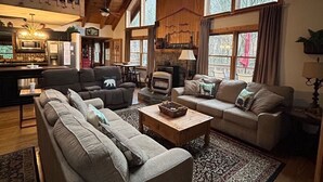 A spacious living room with high ceilings, a ceiling fan, and large windows. There is a fireplace, a sofa set, and a coffee table.