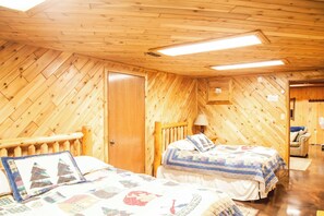 A room with wooden walls and ceiling, two beds with quilts, a wooden door, and a lamp. - Hiawatha’s Hideaway Northwoods Cabin Near Pictured Rocks & Area Lakes (Wetmore)