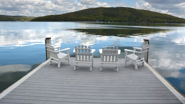Un muelle de madera con tres sillas Adirondack blancas mirando a un lago tranquilo con una colina boscosa al fondo.