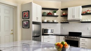 Una cocina moderna con armarios blancos, un refrigerador de acero inoxidable, un microondas y una estufa. Hay una encimera de mármol con un cuenco de fruta.