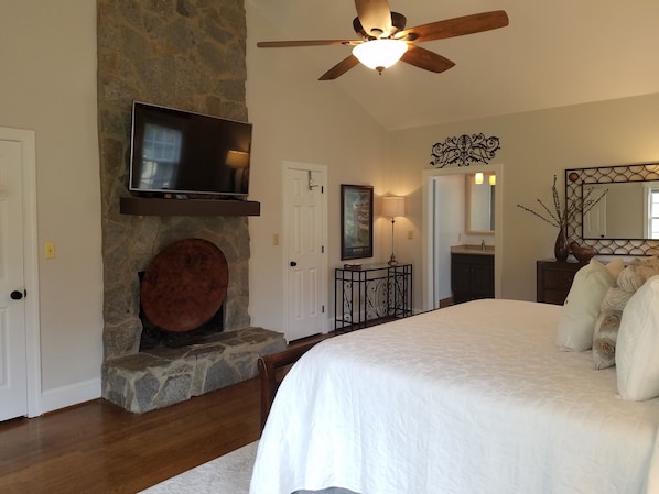 A bedroom with a stone fireplace, a ceiling fan, a TV mounted above it, and a large bed with pillows.