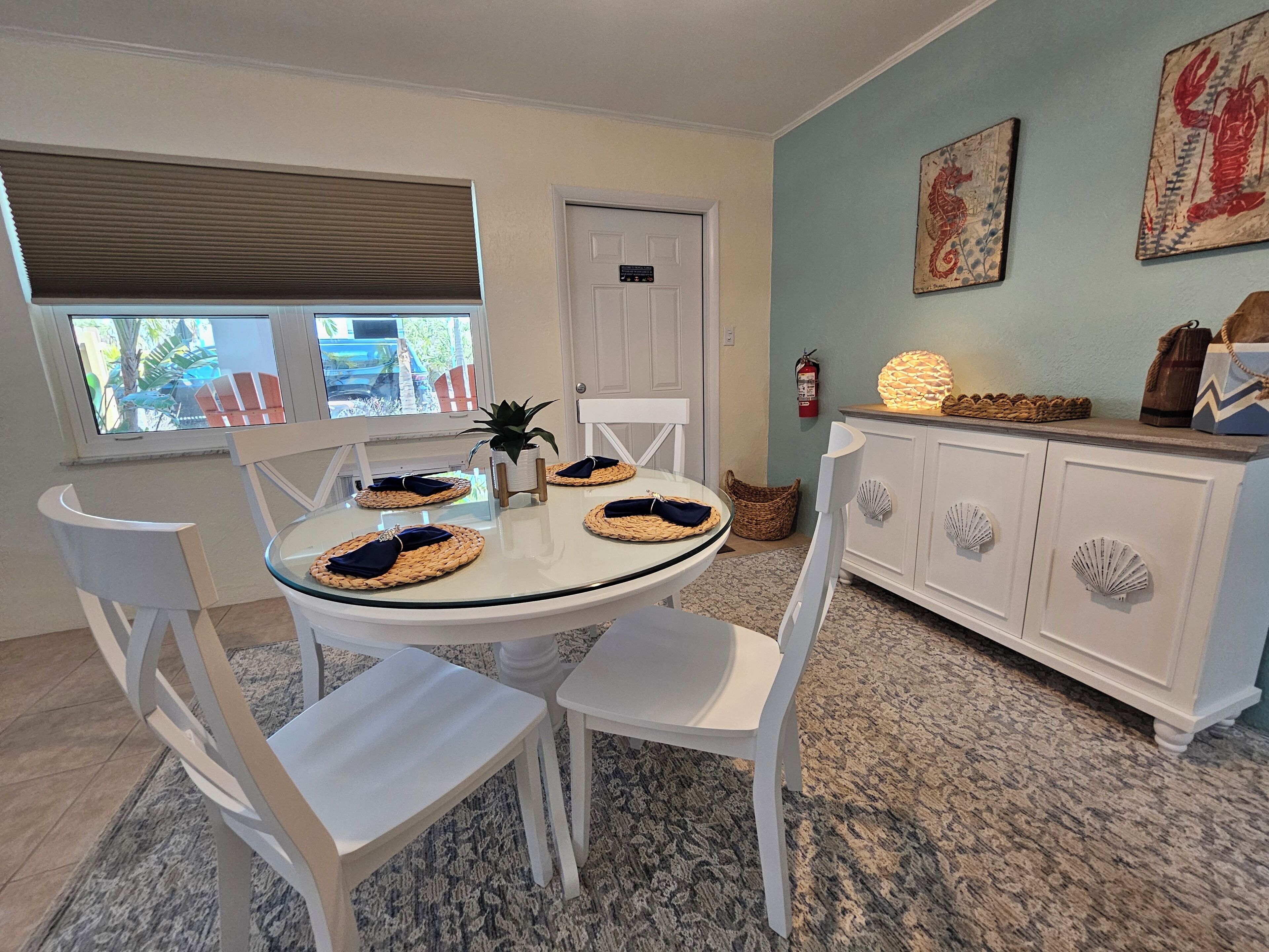 A dining area with a round table set for four, white chairs, a white cabinet with decorative handles, and a window with blinds.