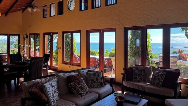 A spacious living area with a large sofa and armchairs, a dining table, and multiple windows offering ocean views. - Amazing Whitewater View Of The Ballena Marine Preserve Islands (Uvita)
