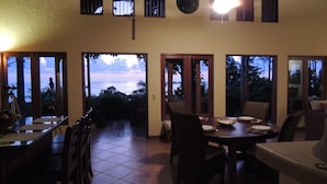 A dining area with a view of the sky through large windows, a ceiling fan, and a dining table set for four. - Amazing Whitewater View Of The Ballena Marine Preserve Islands (Uvita)
