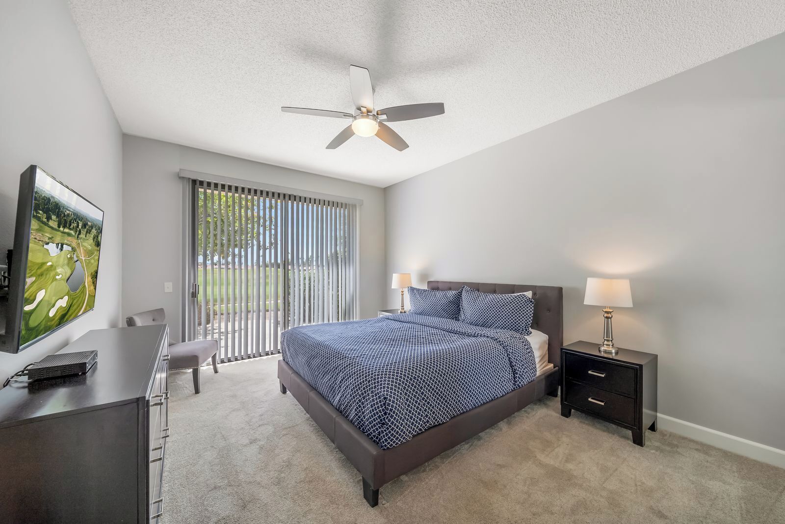 A bedroom with a bed, a desk, a chair, a TV, a ceiling fan, and a window with blinds.