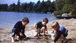Three children playing on a sandy beach near a lake with trees in the background.