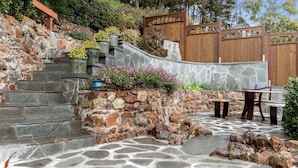 Una escalera de piedra que sube a una terraza de madera con una cascada decorativa, rodeada de macetas con plantas y un suelo de piedra.