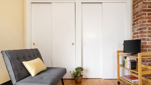 Una habitación con un sofá gris, una pequeña planta en maceta, una estantería con libros y un televisor sobre un mueble de madera.