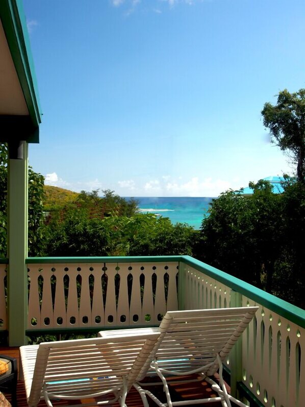 A balcony with two white rocking chairs facing a view of the ocean and lush greenery.