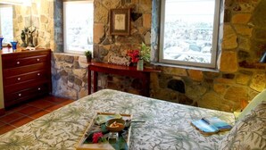 A bedroom with stone walls, a wooden dresser, a bed with a floral bedspread, a window with a view, and a small table with decorative items.