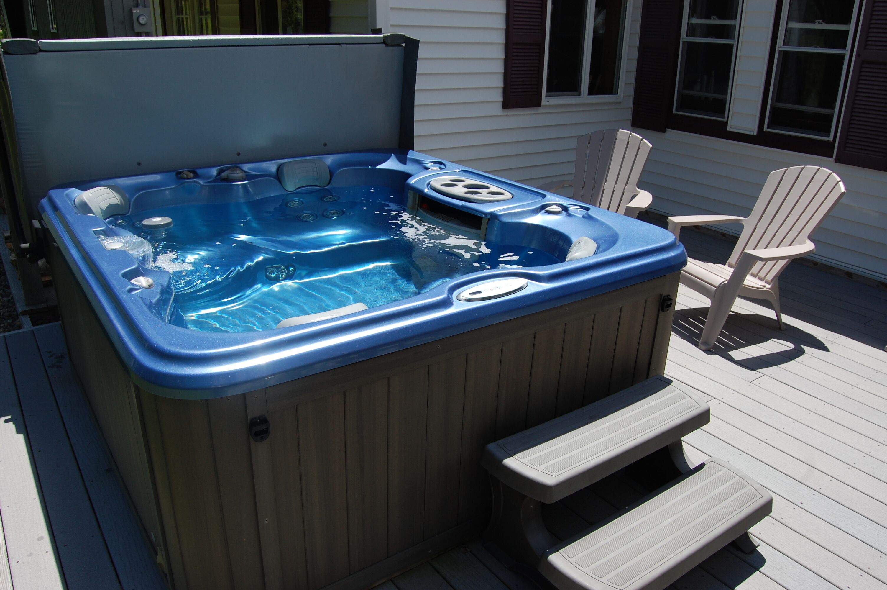 A hot tub with steps and a table, surrounded by Adirondack chairs on a wooden deck.