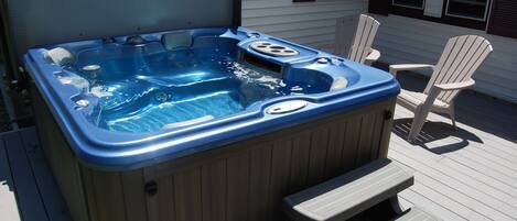 A hot tub with steps and a table, surrounded by Adirondack chairs on a wooden deck.