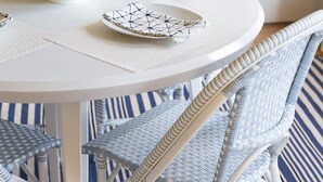 A dining table with a white bowl containing green apples, a white plate with a geometric pattern, and a matching placemat. The table is surrounded by wicker chairs with blue and white stripes, set on a blue and white striped rug.