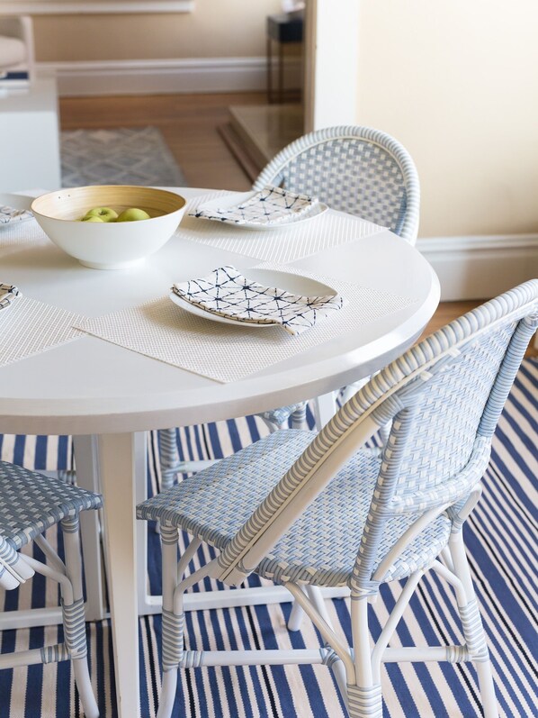 A dining table with a white bowl containing green apples, a white plate with a geometric pattern, and a matching placemat. The table is surrounded by wicker chairs with blue and white stripes, set on a blue and white striped rug.
