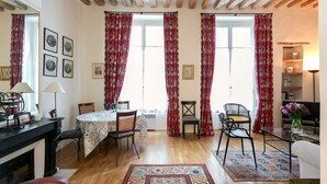 A cozy living room with a fireplace, wooden beams, a dining area with a table and chairs, and large windows with red curtains.