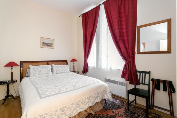 Un dormitorio con cama, una silla, un espejo, un radiador y una ventana con cortinas.