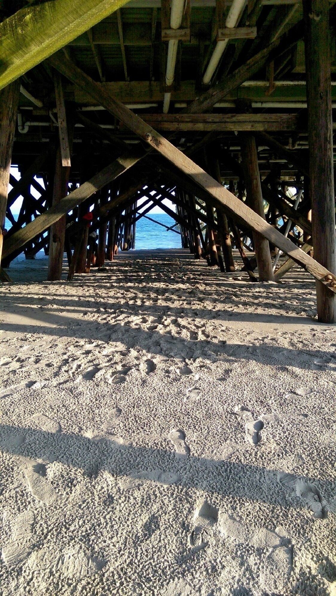 Une plage située sous une jetée en bois, avec des poutres apparentes et une surface sablonneuse.