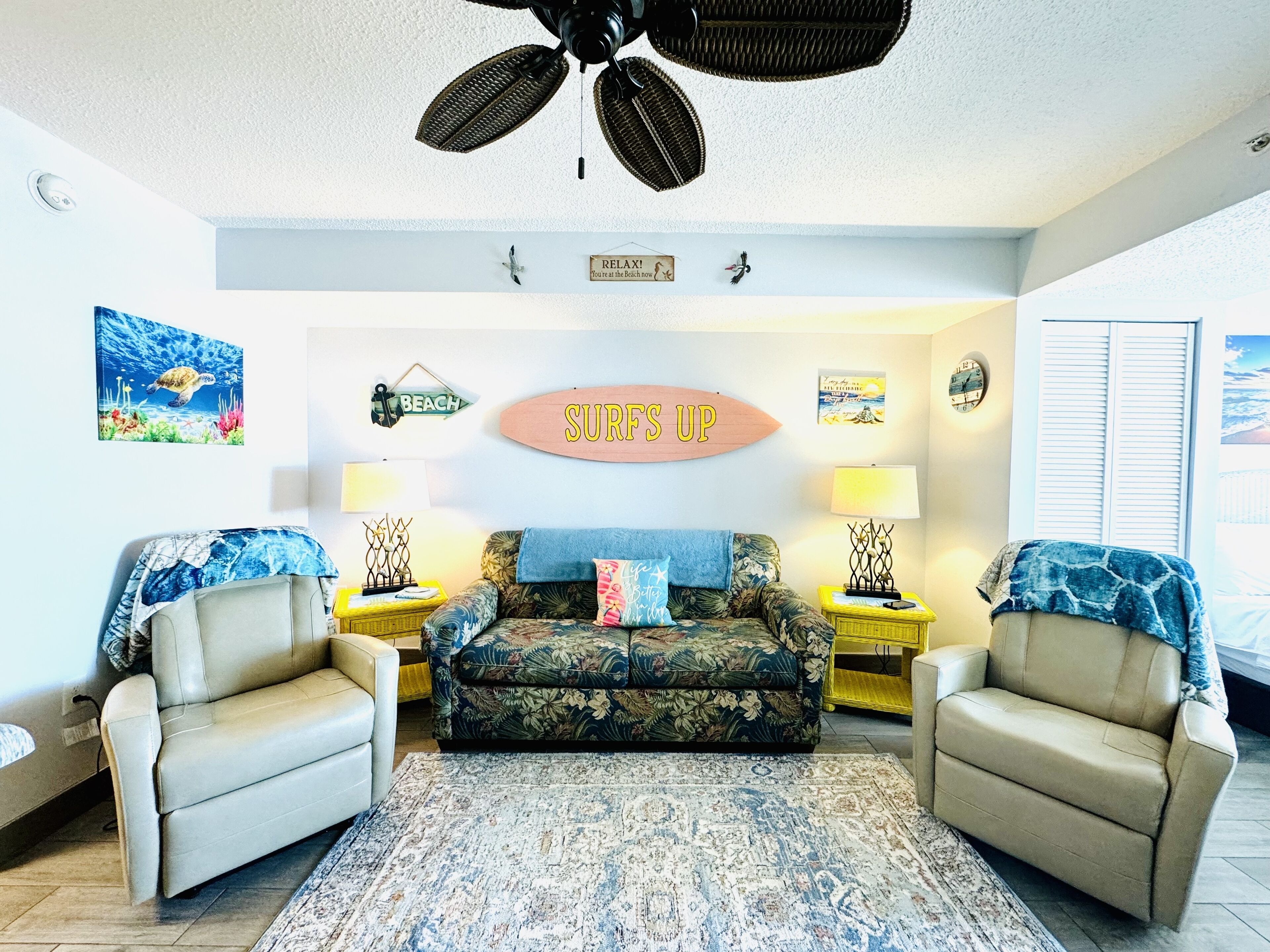 Un salon comprenant un canapé, deux fauteuils, un ventilateur de plafond et des éléments de décoration murale, notamment un panneau représentant une planche de surf.
