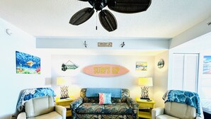 A living room with a sofa, two armchairs, a ceiling fan, and wall decorations including a surfboard sign. - 💎NEWLY REMODELED!! ISLAND TIME Honeymoon Suite Direct Oceanfront King Luxury (Myrtle Beach)