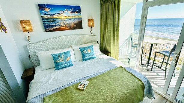 A hotel room with a bed, a painting of a sunset, a balcony with a view of the ocean, and a small table with a chair. - 💎NEWLY REMODELED!! ISLAND TIME Honeymoon Suite Direct Oceanfront King Luxury (Myrtle Beach)