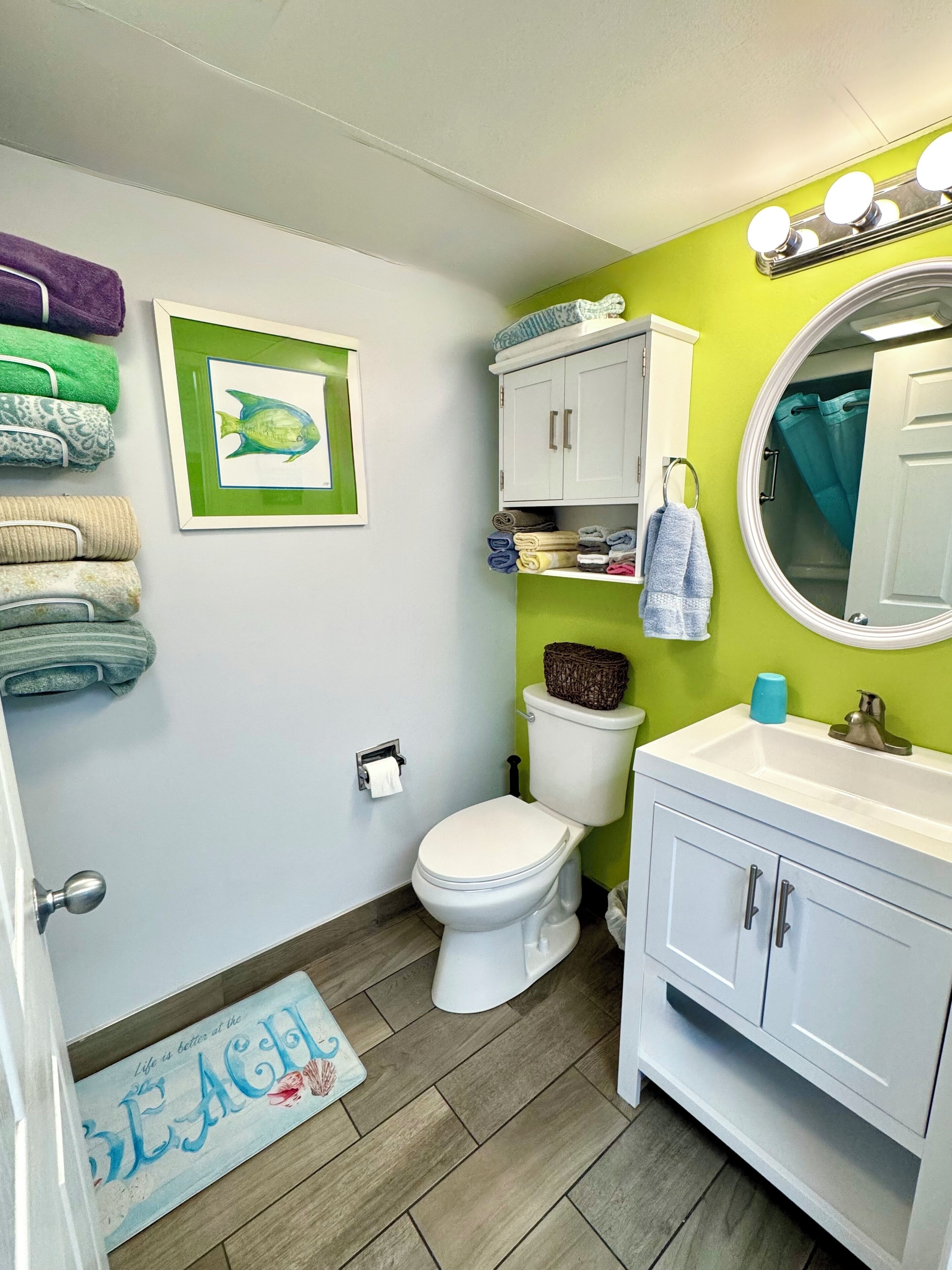 A bathroom with a toilet, sink, and a towel rack with towels. There is a framed picture on the wall and a beach-themed rug on the floor.