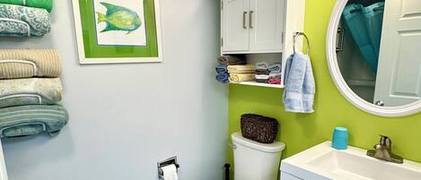 A bathroom with a toilet, sink, and a towel rack with towels. There is a framed picture on the wall and a beach-themed rug on the floor.