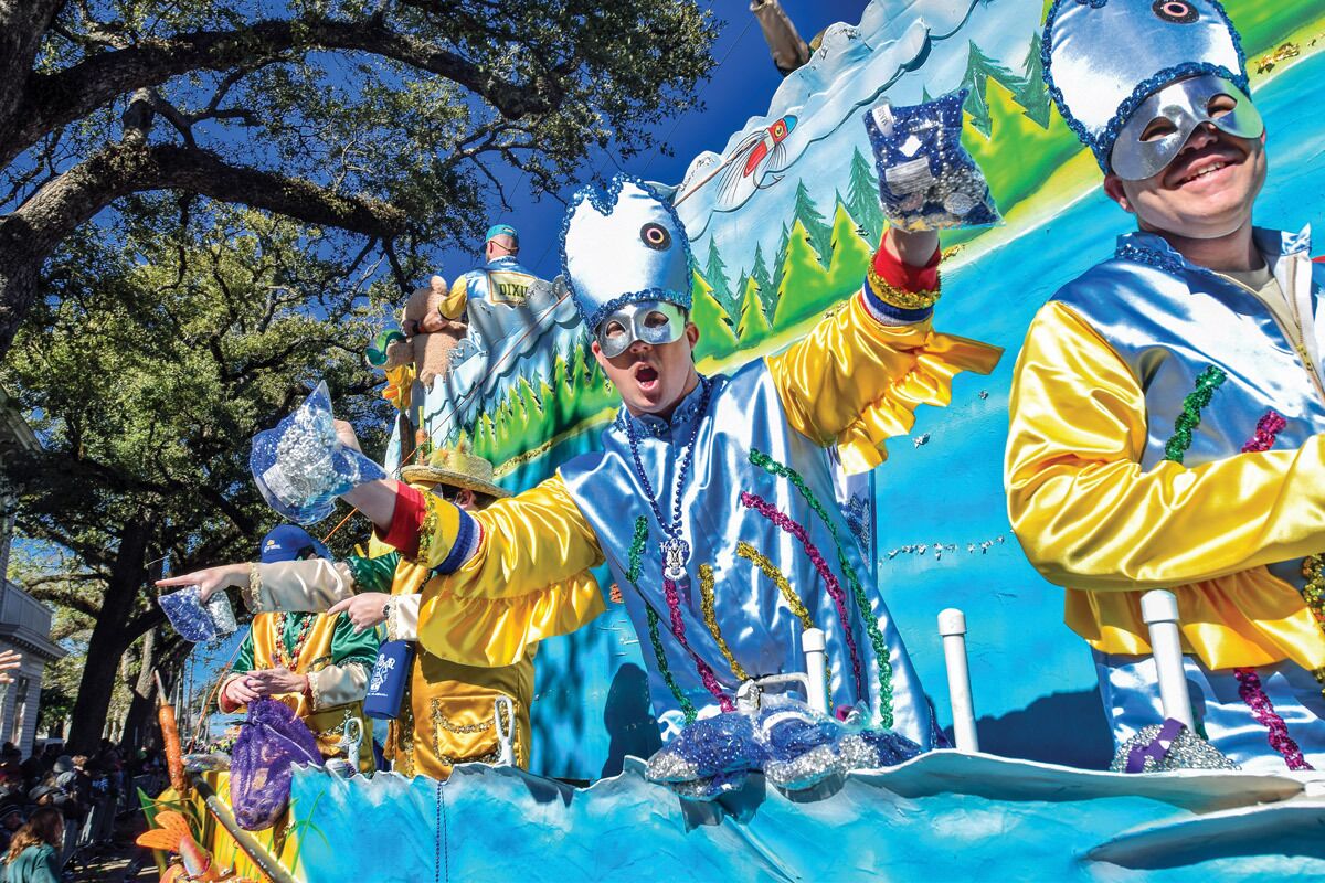 Un carroza de desfile con artistas vestidos con trajes y máscaras coloridos, sobre un gran fondo azul que muestra árboles y un río.