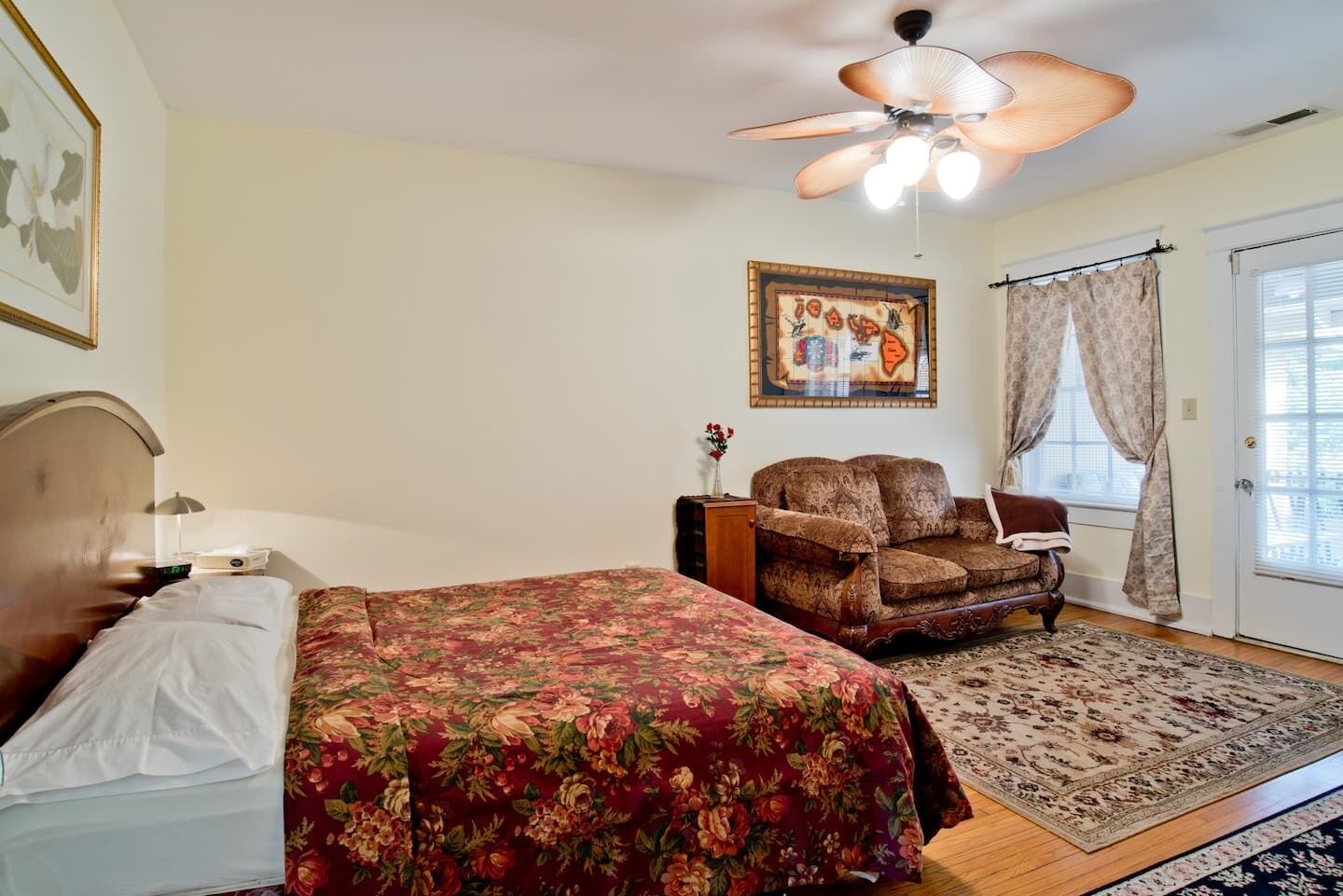 A bedroom with a bed, a chair, a ceiling fan, a painting, and a door with curtains.