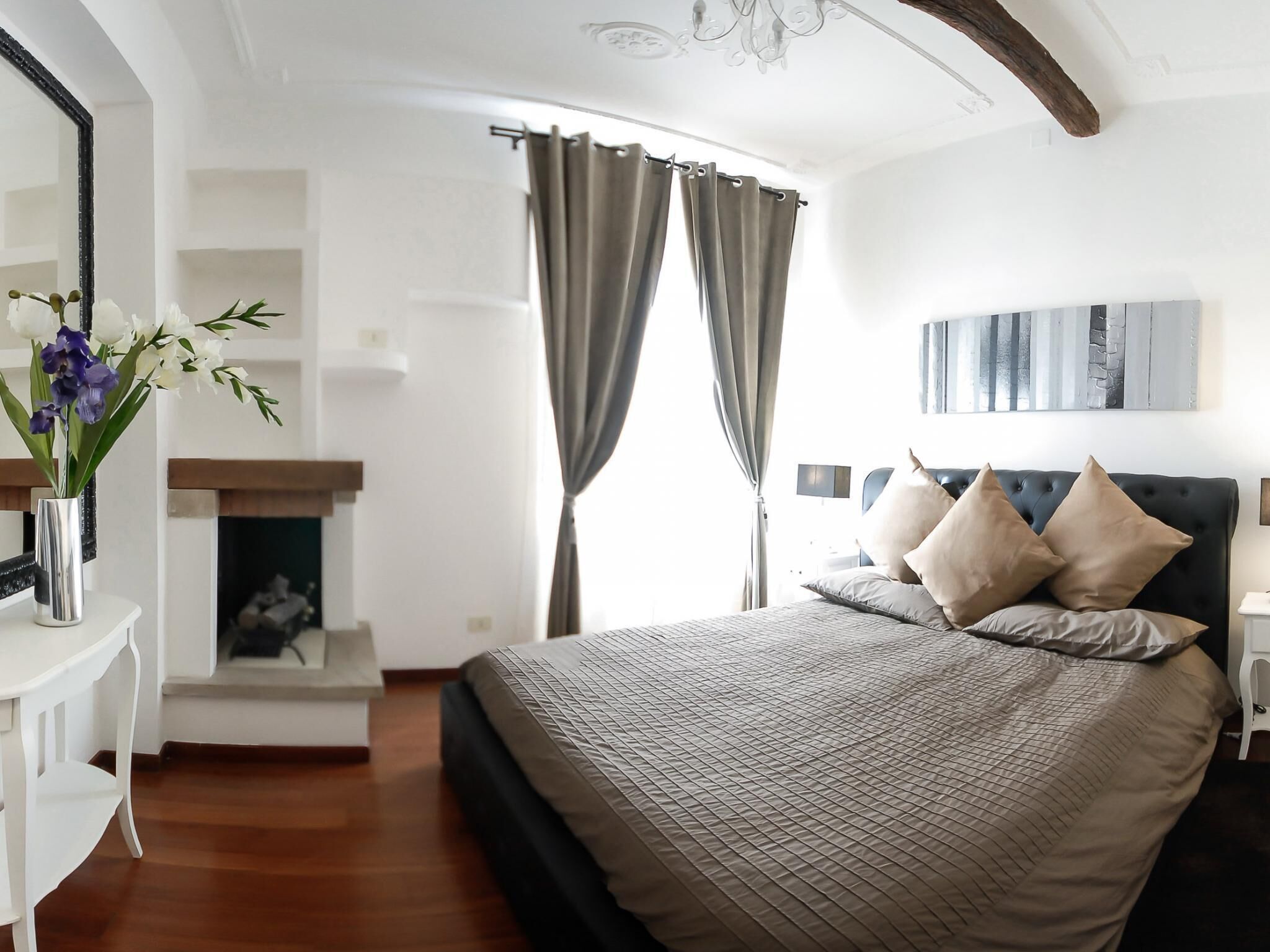 A bedroom with a bed, a nightstand, a mirror, a fireplace, and a painting on the wall.