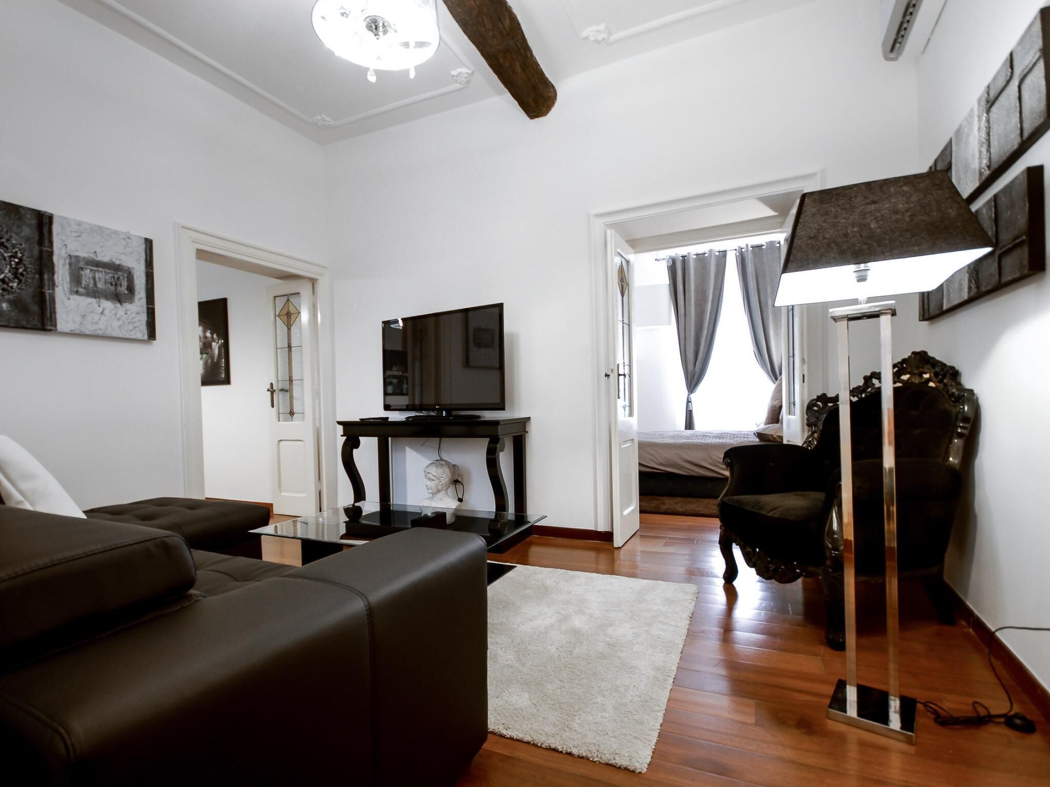 A modern living room with a sofa, a TV on a stand, a lamp, and a bed in the background.