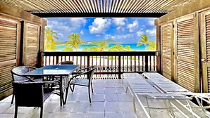 Un espace extérieur abrité, avec une table et des chaises, offrant une vue sur une plage et des palmiers, et doté d’une structure de plafond en bois.