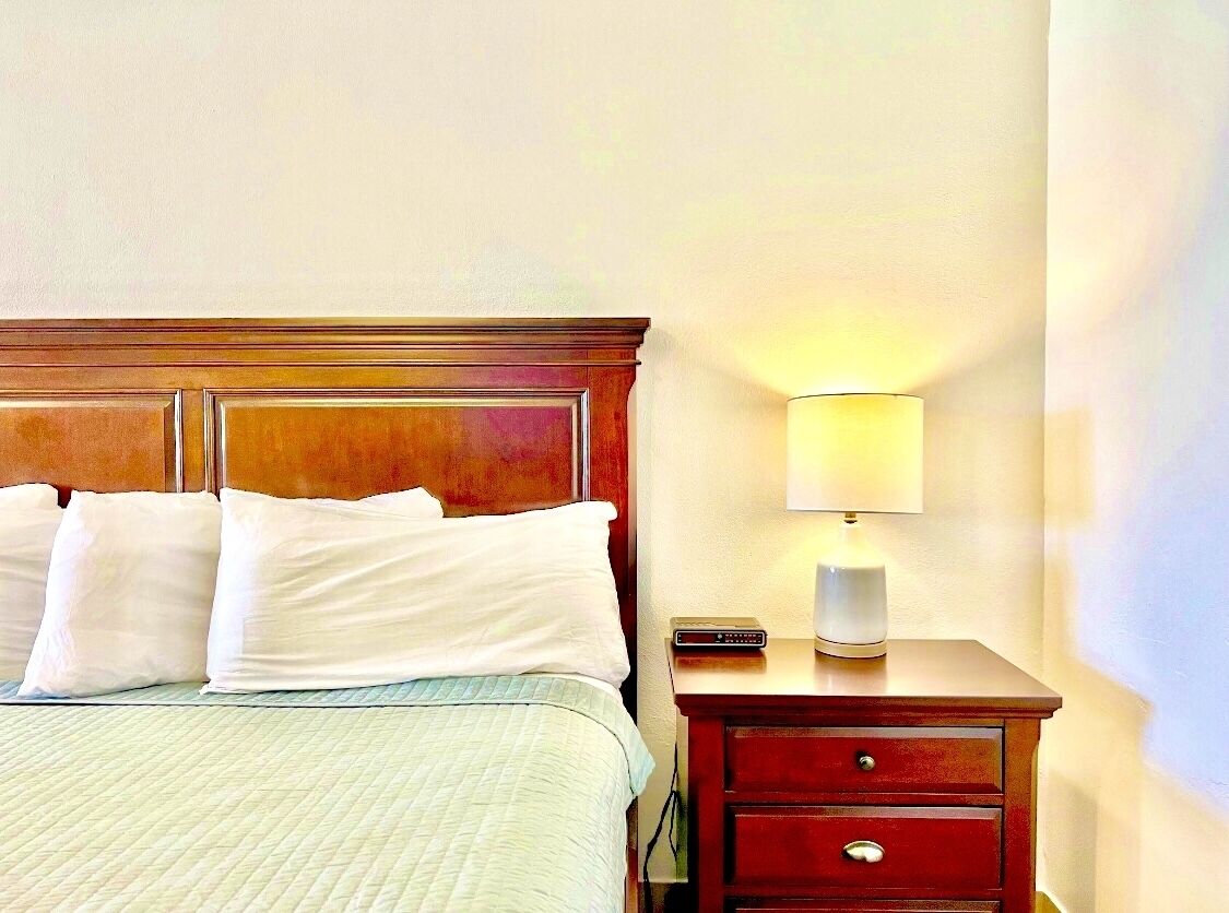 A hotel room with a bed, pillows, a bedside table with a lamp, and a remote control.