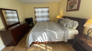 Un dormitorio con cama, cómoda, televisor, mesita de noche con lámpara y un cuadro en la pared.