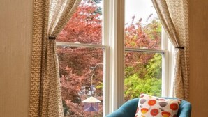 Un sillón color turquesa con un cojín floral, una mesa de centro de madera con un florero y una ventana con cortinas estampadas.