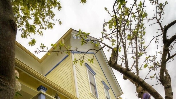 Una casa amarilla de dos pisos con un balcón y flores púrpuras en primer plano.