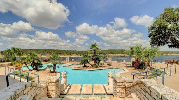 A swimming pool area with a stone wall, palm trees, and a view of the surrounding landscape. - Splendid, Tastful, And Fully Furnished Condo On Lake Travis Island Villa!!! (Lago Vista)
