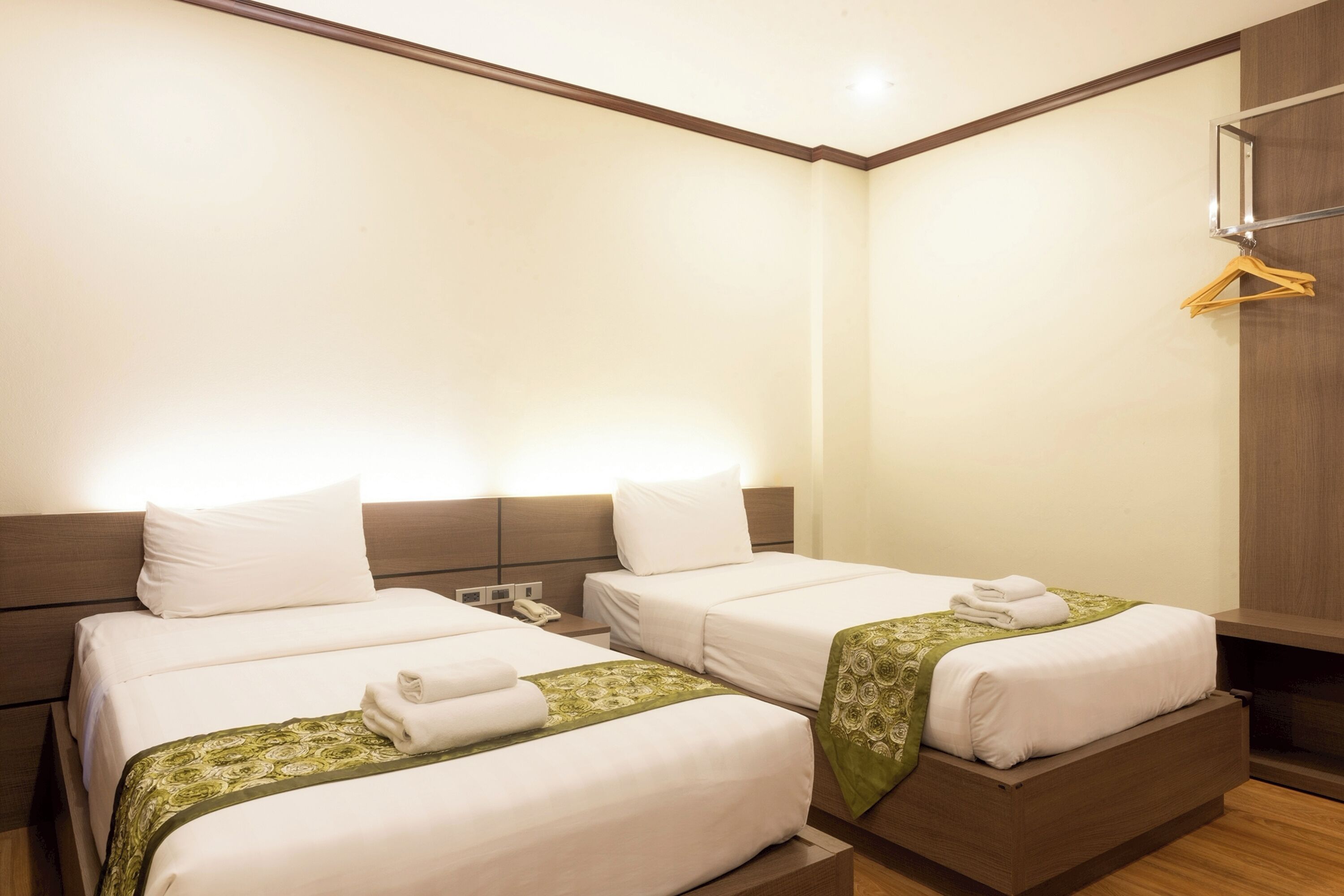 A hotel room with two beds, a wooden headboard, a bedside table with a phone, and a wall-mounted hanger.