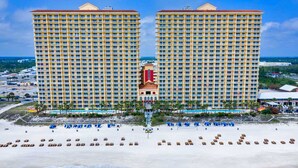 Ein großes Hotel mit mehreren Etagen und Balkonen, einem zentralen Turm sowie einem Strandbereich mit Liegestühlen und Sonnenschirmen.