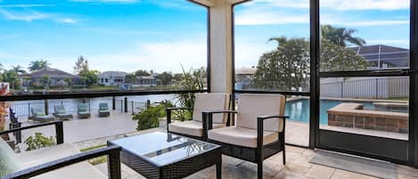 A balcony with wicker furniture, a glass table, and a view of a swimming pool and houses.