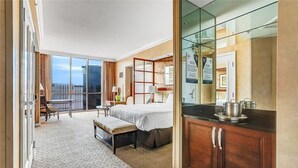 Ein Hotelzimmer mit Bett, Schreibtisch, Stuhl, kleinem Tisch, Bank, Küchenzeile inklusive Herd und Spüle sowie einem großen Fenster mit Blick auf Gebäude.