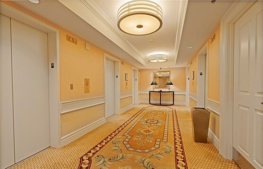 A hotel hallway with a patterned carpet, white doors, and a central light fixture.