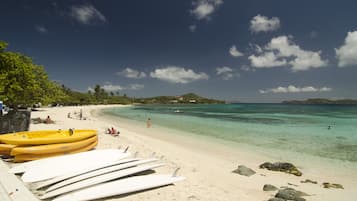 Una playa con tablas de surf y kayaks, agua turquesa cristalina y algunas personas a lo lejos.