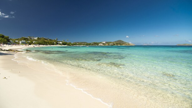 Ein Strand mit klarem türkisblauem Wasser, sandigem Ufer und Gebäuden im Hintergrund.